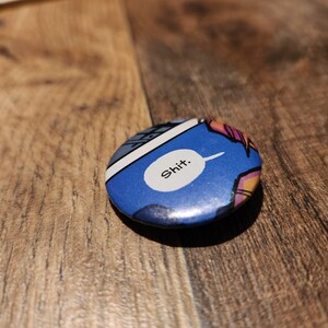 May include: A round button with a comic book style design. The button has a blue background with a white speech bubble containing the word "Shit." The button has a glossy finish and is set against a wooden surface.