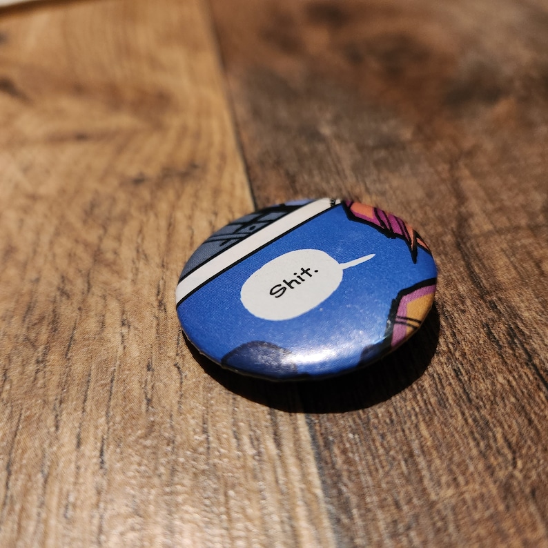 May include: A round button with a comic book style design. The button has a blue background with a white speech bubble containing the word "Shit." The button has a glossy finish and is set against a wooden surface.