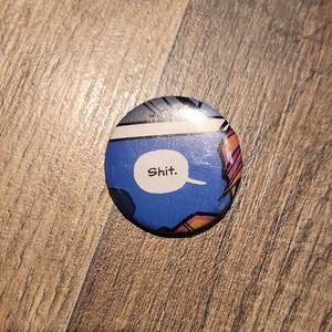 May include: Round button with a comic book panel design. The button features a blue background with a white speech bubble containing the word "Shit." The button has a glossy finish and is set against a wooden surface.