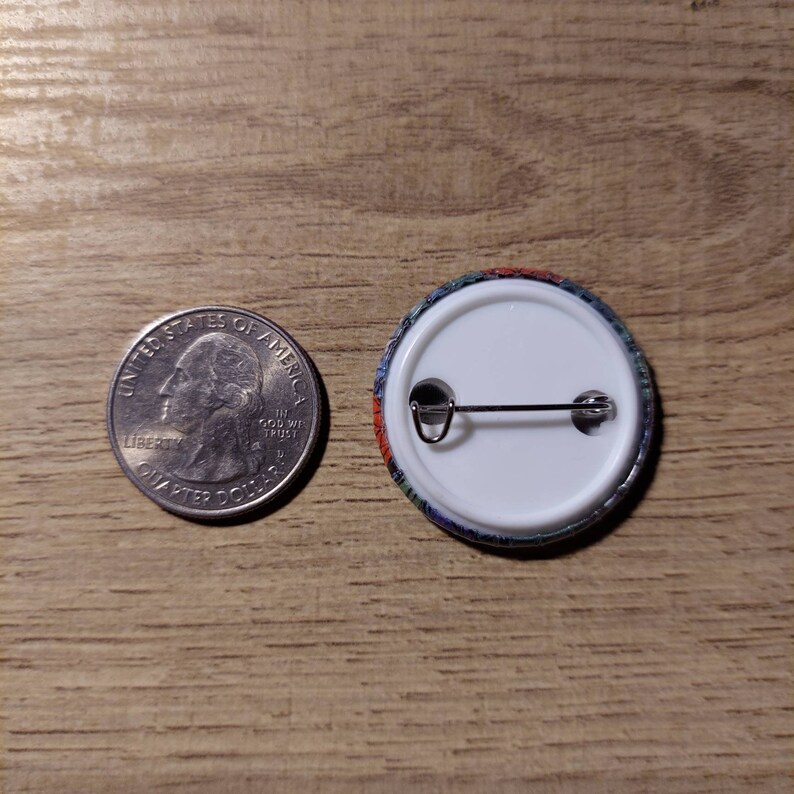 May include: A round button with a colorful design, next to a U.S. quarter. The button has a white back with a pin. The button's design includes red, blue, and green colors. The button is approximately 1 inch in diameter.