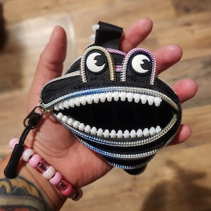 May include: A black zippered pouch with a zippered mouth that resembles a toothy grin. The pouch has two large white eyes with black pupils and a rainbow colored zipper.
