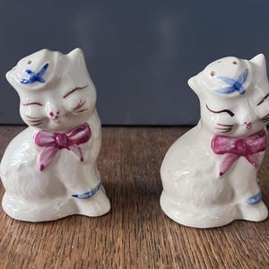 Może przedstawiać: Para ceramicznych solniczek i pieprzniczek w kształcie kota w stylu vintage. Białe koty mają zamknięte oczy, różowe kokardy i niebieskie akcenty. Solniczki stoją na drewnianej powierzchni.