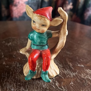 Peut inclure: Figurine d'elfe en céramique avec un chapeau et un pantalon rouges, une chemise verte et des bottes vertes. L'elfe est assis sur une branche d'arbre. La figurine a un visage souriant et est peinte avec des couleurs vives.
