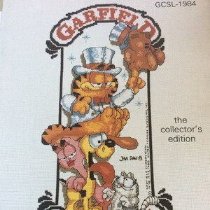 Può includere: Un modello a punto croce con i personaggi di Garfield and Company. Garfield, con un cappello a cilindro, è in cima, con Odie e altri personaggi sotto. Il testo "GARFIELD AND COMPANY" è in basso, con il testo "the collector's edition".