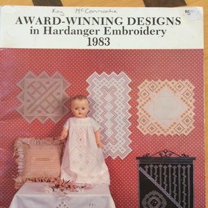 Peut inclure: Couverture de livre "AWARD-WINNING DESIGNS in Hardanger Embroidery 1983" avec une poupée en robe blanche et des exemples de broderie Hardanger. Le fond est rouge à pois blancs. La couverture est sur une table avec une nappe blanche.