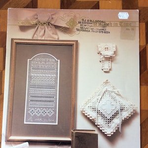 May include: An image of a cross-stitch pattern book and related items. The book cover features a framed cross-stitch sampler with the alphabet. Other items include a needlework book, a pincushion, and a decorative pillow.