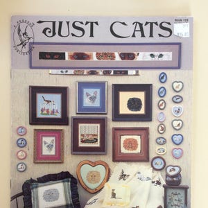 Puede incluir: La portada del libro "JUST CATS" presenta varios diseños de punto de cruz con temática de gatos. Los diseños incluyen imágenes enmarcadas, marcos en forma de corazón y pequeños objetos decorativos, todos mostrando diferentes razas y estilos de gatos.