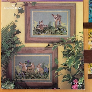 Può includere: Un opuscolo di modelli di ricamo a punto croce con due immagini incorniciate di animali della foresta. L'immagine superiore mostra due cervi in un ambiente boschivo. L'immagine inferiore mostra tre conigli in un ambiente boschivo. La copertina dell'opuscolo è viola con il titolo "Forest Friends" e il testo "Anniversary Collection 10 Years of Just Cross Stitch" in un cerchio. Il testo "by Charlotte Holder" è sotto il titolo. Il testo "Item #2014 5,00 €" è nell'angolo in basso a destra.