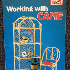 Puede incluir: Portada de libro vintage "Working with Cane" con una estantería y una silla de bambú sobre un fondo azul. La estantería tiene estantes de vidrio y está decorada con frutas, flores y una planta. La silla tiene un cojín rojo.