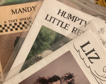 Może przedstawiać: Stosik zabytkowych broszur z wzorami lalek. Okładki zawierają tytuły takie jak "Mandy" i "Humpty Little Red". Broszura "Liz" przedstawia dwie ręcznie robione lalki, jedną w szachownicowym stroju, a drugą w czerwono-niebieskim.