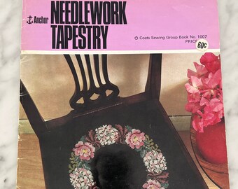May include: A vintage "Needlework Tapestry" book cover featuring a dark wooden chair with a needlepoint seat. The seat displays a floral wreath design in pink, white, and green. The book cover is pink with black text and the Anchor logo.