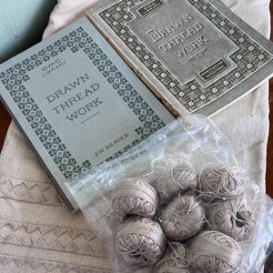 Peut inclure: Deux livres anciens intitulés "Drawn Thread Work" de la série DMC Library, avec des couvertures grises et des bordures décoratives. Un sac en plastique transparent contient plusieurs bobines de fil beige. Un tissu brodé beige est également visible.