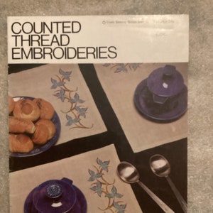 May include: A book cover with the title "Counted Thread Embroideries". The cover features a photo of two embroidered placemats with a blue and white floral design. The placemats are set on a dark surface with a blue and white ceramic dish and spoons.