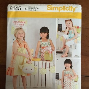 Puede incluir: Un patrón de costura Simplicity para vestidos de niñas. El patrón incluye tres estilos diferentes: un vestido amarillo con un dobladillo con volantes, un vestido de lunares rojos y blancos y un vestido de rayas azules y blancas. El patrón es para las tallas 3-8.