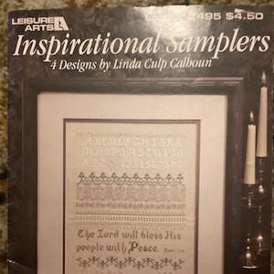 Op de afbeelding: Een ingelijst borduurwerk met de tekst "The Lord will bless His people with Peace." en het alfabet en de cijfers. Het borduurwerk is in een bruin frame en ligt op een tafel met een boek.