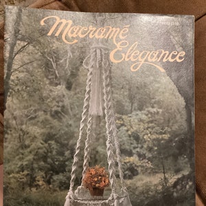 May include: A vintage book cover titled "Macramé Elegance" featuring a white macramé plant hanger with a pot of orange flowers. The plant hanger has a round base and is suspended in front of a blurred forest background. The text "La Creation" is at the bottom.