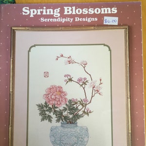 以下が含まれることがあります： Serendipity Designsの「Spring Blossoms」というタイトルの額入りクロスステッチパターン。ピンクの牡丹と繊細なピンクの花が青い花瓶に描かれています。フレームは金色で、背景はソフトピンクです。