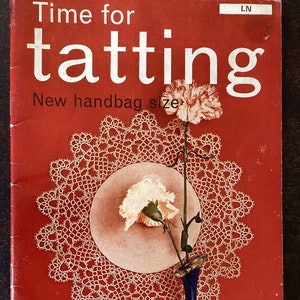 Puede incluir: Una portada de libro vintage con el título "Time for tatting" y el subtítulo "New handbag size". La portada presenta un encaje blanco hecho a mano, tres claveles rosas en un jarrón azul y un fondo rojo.