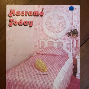 May include: A pink and white macrame bedspread with a white crocheted wall hanging and a pink and white patterned wallpaper. The text on the cover reads "Macrame Today" and "Exciting New Designs Including Macra-Weaving and Macra-Sculpture".