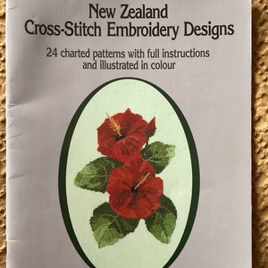 May include: Book cover featuring a cross-stitch embroidery design of two red hibiscus flowers with green leaves. The title is "New Zealand Cross-Stitch Embroidery Designs" and the subtitle is "24 charted patterns with full instructions and illustrated in colour". The author is Margaret Barrett.
