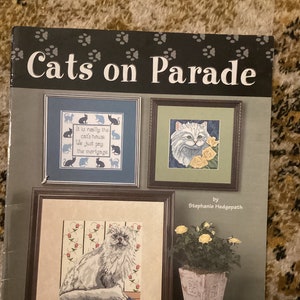 Puede incluir: Patrón de punto de cruz con un gato blanco con flores amarillas. El patrón se titula "Cats on Parade" e incluye el texto "It is really the cat's house. We just pay the mortgage." El patrón es de Stephanie Hedgepath y está disponible en Jeanette Crews Designs.