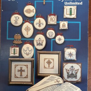 May include: A book cover titled "Christian Symbols Unlimited" with various cross-stitch designs of Christian symbols. The designs include crosses, fish, crowns, and other religious icons. The book is open to a page with text and a bookmark.