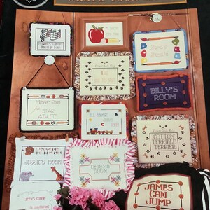 Può includere: Una collezione di schemi a punto croce con vari cartelli per stanze. I disegni includono "What's Your Sign?", "Michael's Room", "Billy's Room" e "James Can Jump". I cartelli sono incorniciati e decorati con vari motivi.