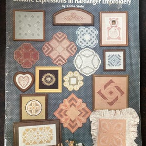May include: A softcover book titled Creative Expressions In Hardanger Embroidery by Zetha Stuhr, featuring a blue polka-dot cover displaying various framed and unframed Hardanger embroidery projects in white, pink, brown, and gold thread.