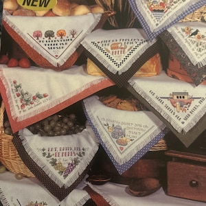 Könnte beinhalten: Eine Sammlung weißer Brottücher mit ausgefransten Rändern, die jeweils mit Kreuzstichmotiven und religiösen Schrifttexten versehen sind. Die Tücher sind über verschiedene Körbe und Behälter drapiert und zeigen farbenfrohe Motive wie Bäume, Weizen, Obst und Brot.