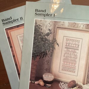 Könnte beinhalten: Zwei Stickmuster-Vorlagen, eine mit dem Titel "Band Sampler I" und die andere mit "Band Sampler II". Das Muster für "Band Sampler I" zeigt ein gerahmtes Stickmuster mit dem Text "She seeks wool & flax & works with willing hands".