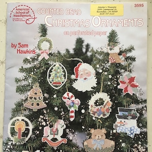 Può includere: Una collezione di ornamenti natalizi a perline contate su carta perforata. Gli ornamenti includono un albero di Natale, Babbo Natale, un pupazzo di neve, un orsacchiotto e altri disegni festivi. Il testo sulla copertina recita "American School of Needlework" e "Christmas Ornaments."