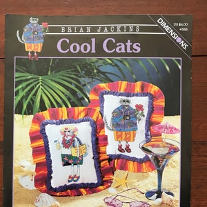 May include: Cool Cats cross stitch pattern by Brian Jackins. The pattern features two cats wearing Hawaiian shirts and sunglasses. The pattern is from Dimensions, and the pattern number is 359. The pattern is on a white background with a beach scene.