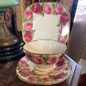 Puede incluir: Un juego de taza y platillo de té vintage con un patrón floral. La taza y el platillo son blancos con un borde dorado y presentan rosas rosadas con hojas verdes.