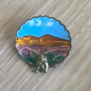 Pode incluir: Um broche de esmalte redondo com uma cena de paisagem. O broche apresenta um céu azul com nuvens brancas, uma cordilheira marrom e colinas roxas em primeiro plano. Uma pequena figura está sentada em primeiro plano.