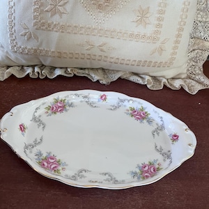 Puede incluir: Un plato ovalado para servir con fondo blanco y borde dorado. El plato está decorado con un patrón floral de rosas rosas y hojas grises.