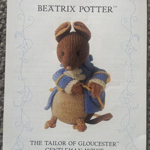 Puede incluir: Una figura de ratón de punto de "The World of Beatrix Potter". El ratón lleva un abrigo azul con ribetes dorados y un chaleco beige. El texto "The Tailor of Gloucester Gentleman Mouse" es visible.