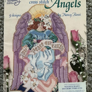Può includere: Un libro di schemi a punto croce intitolato "Angels" di Nancy Rossi. La copertina presenta un angelo a punto croce con ali viola, che tiene uno striscione con la scritta "You are not alone on earth". Il libro include 9 disegni.