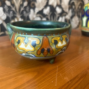 May include: A small ceramic bowl with a green interior and three small feet. The exterior has a decorative band with orange, yellow, and blue floral patterns. The bowl is likely vintage or antique.