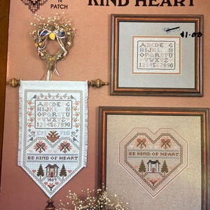 May include: An image featuring cross-stitch samplers and a heart-shaped wreath. The samplers include framed pieces and a banner, all with the text "KIND HEART" and cross-stitch patterns. The banner reads "BE KIND OF HEART".