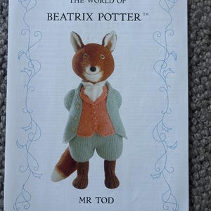 Puede incluir: Un papel impreso con el texto The World of Beatrix Potter en la parte superior y Mr. Tod en la inferior. En el centro, una figura de zorro tejida lleva una chaqueta verde claro, un chaleco naranja, un pañuelo blanco y pantalones verde claro.