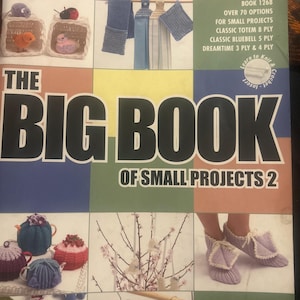 May include: A softcover craft book titled The Big Book of Small Projects 2 by Patons, featuring a grid of various knitted and crocheted items including tea cozies, slippers, hanging ornaments, and small toys against a colorful background.