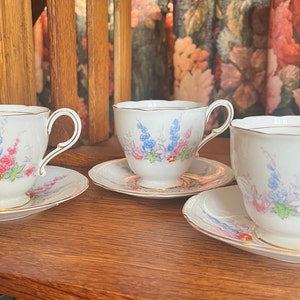 May include: Three white teacups and saucers with a floral pattern in shades of blue, pink, and green. The cups have a gold rim and the saucers have a gold band around the edge.