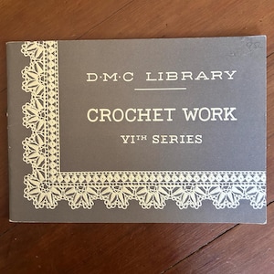 May include: A rectangular gray booklet titled D.M.C LIBRARY CROCHET WORK VIth SERIES in white text. The cover features a decorative cream-colored lace border pattern along the left and bottom edges, printed on a matte gray background.