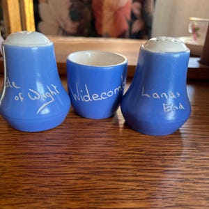 Puede incluir: Tres saleros y pimenteros de cerámica azul con letras blancas. Los saleros están etiquetados como "Isle of Wight", "Widecombe" y "Land's End".