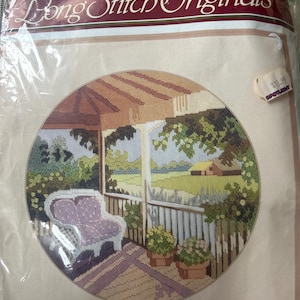 Puede incluir: Un kit de bordado Semco Long Stitch Originals titulado Shady Verandah, número de kit 3300-3110. El empaque presenta una ilustración circular de un porche con una silla morada, flores amarillas en macetas y una vista de un campo verde.