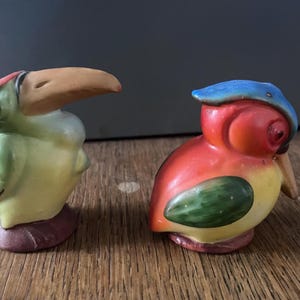 Pode incluir: Duas figuras de pássaros de cerâmica. Um é um tucano com corpo verde, barriga amarela e bico laranja grande. O outro é um papagaio com corpo vermelho, cabeça azul e barriga amarela. Ambos têm um acabamento brilhante.
