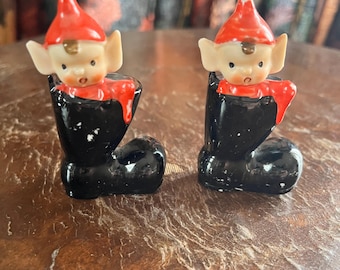 May include: A pair of vintage salt and pepper shakers shaped like elves peeking out of black boots. The elves have red hats and white faces with brown hair.