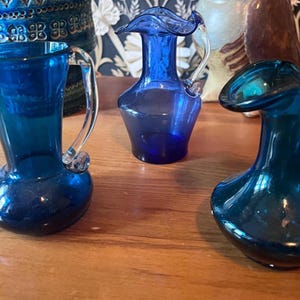 Puede incluir: Tres jarras de vidrio azul cobalto con asas de vidrio transparente. Las jarras tienen una base bulbosa y un borde acampanado. Las jarras se exhiben sobre una superficie de madera. Las jarras son decorativas y adecuadas para servir bebidas.