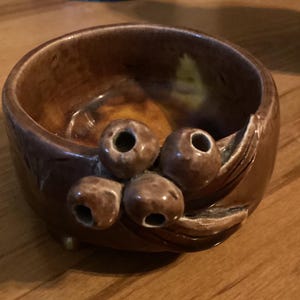 Puede incluir: Un pequeño cuenco de cerámica marrón con una superficie texturizada y un diseño de hojas y bayas. El cuenco tiene un acabado esmaltado y una forma ligeramente redondeada.