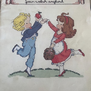 Può includere: Schema a punto croce con due bambini, un ragazzo e una ragazza, in posa giocosa. Il ragazzo, in tuta blu e giacca blu navy, offre una mela rossa alla ragazza, che indossa un vestito rosso e porta un cestino di fiori. Il testo "joan walsh anglund" è sopra.